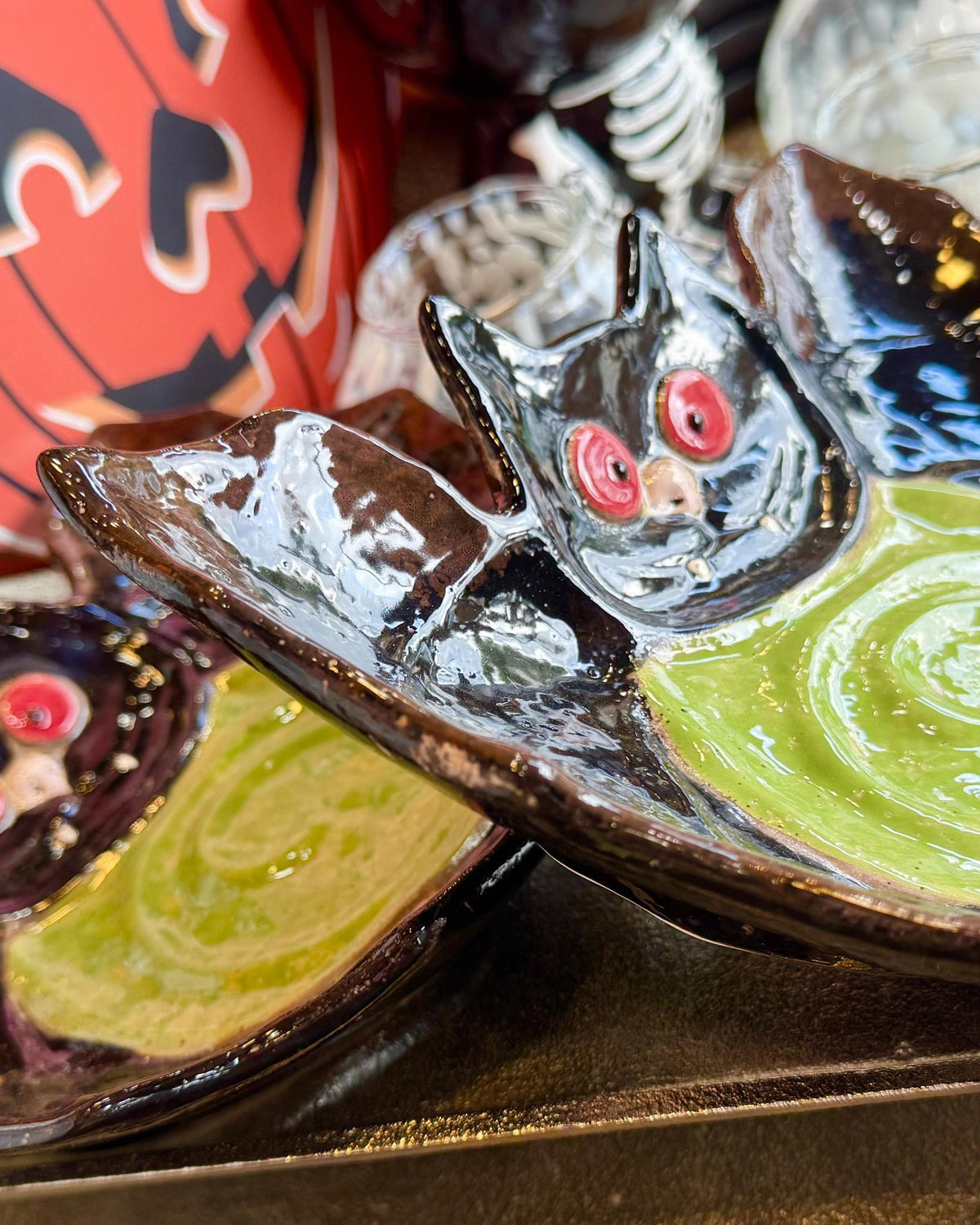 A close-up of a glossy ceramic plate shaped like a bat, featuring a black bat head with red eyes and wings spread over a bright green swirl of paint or glaze, set against a dark tray with other decorative plates.
