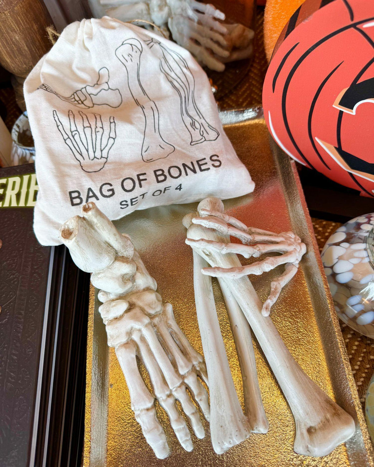 A Halloween prop set featuring a set of four fake human bones and a small drawstring bag labeled 'BAG OF BONES SET OF 4' with foot bone illustrations, displayed on a gold tray.