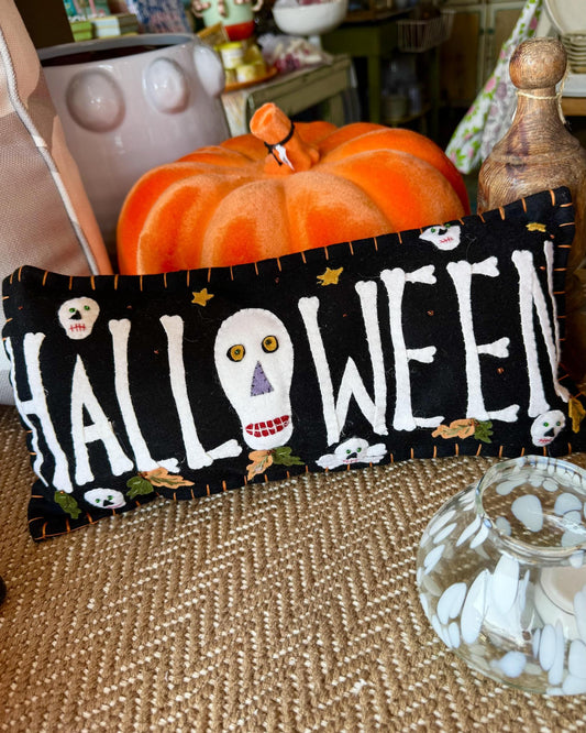 A black fabric Halloween pillow with white embroidered 'HALLOWEEN' lettering, skull and ghost motifs, and small decorative details, styled in a cozy home setting with a pumpkin and woven table runner.