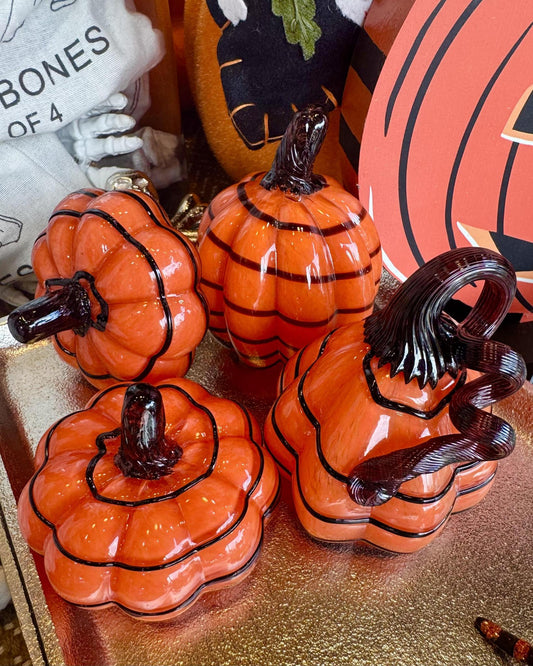 Four glossy orange glass pumpkins with black decorative lines and dark stems, arranged on a gold tray with Halloween-themed decor in the background.