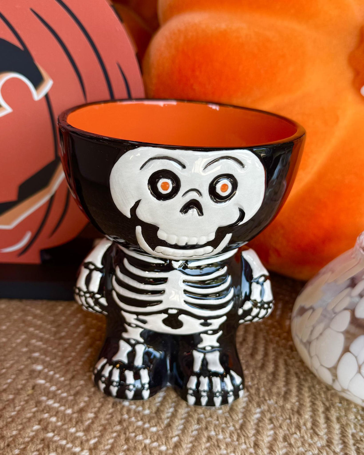 A ceramic skeleton-shaped cup or candle holder with a black exterior, white skeleton design, and bright orange interior, styled with Halloween decorations in the background.