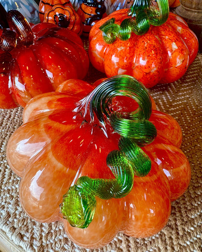 Three decorative hand-blown glass pumpkins in orange, red, and amber tones with green stems, arranged on a woven surface.
