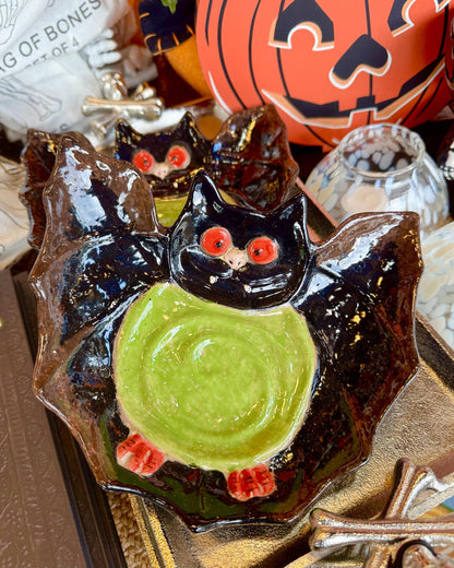 A ceramic Halloween-themed bowl shaped like a black bat with a green circular center and red eyes displayed on a tabletop with seasonal decor in the background.
