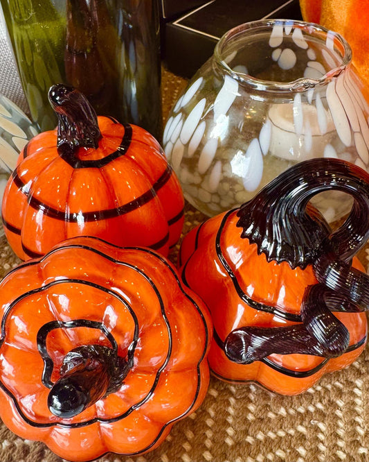 Three orange ceramic pumpkins with black stems and outlines, arranged on a woven surface with glassware in the background.