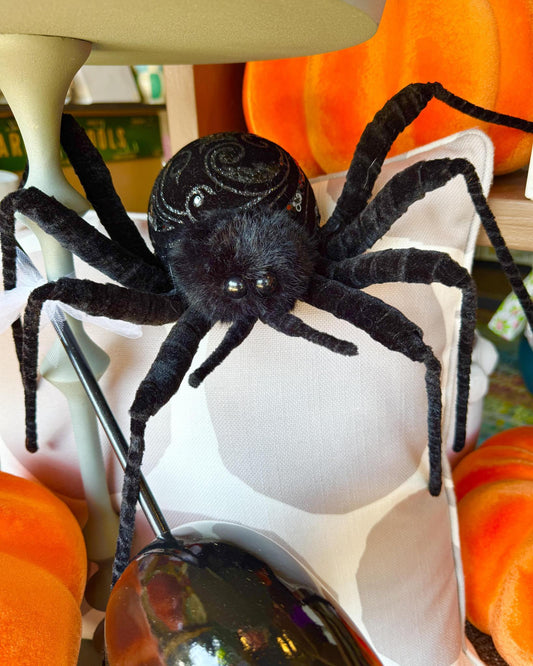 A black plush spider decoration with glittery details on its abdomen, posed on a white pillow with orange pumpkins in the background.