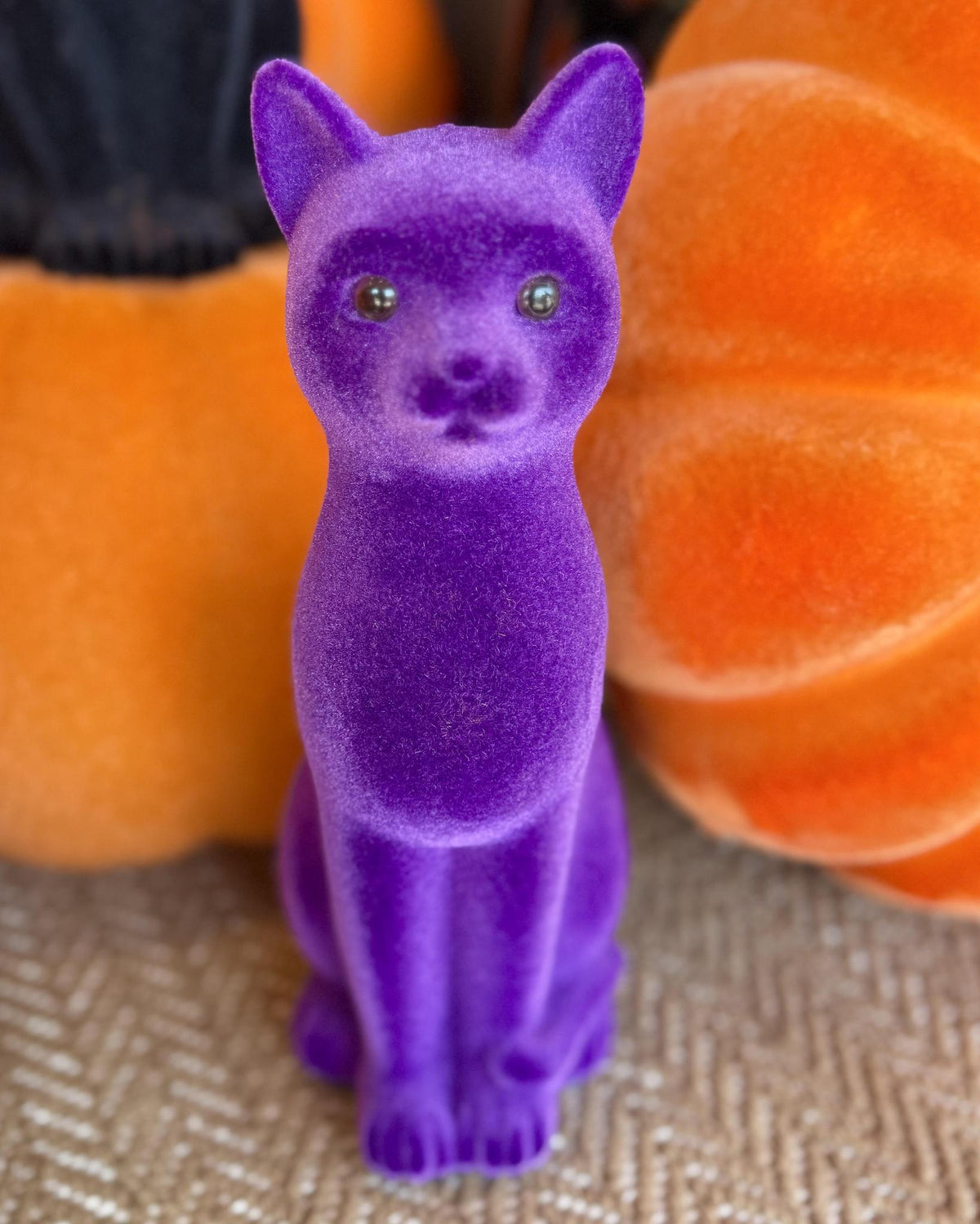 A purple cat figurine with green eyes, standing upright on a textured beige surface with orange pumpkins in the background.