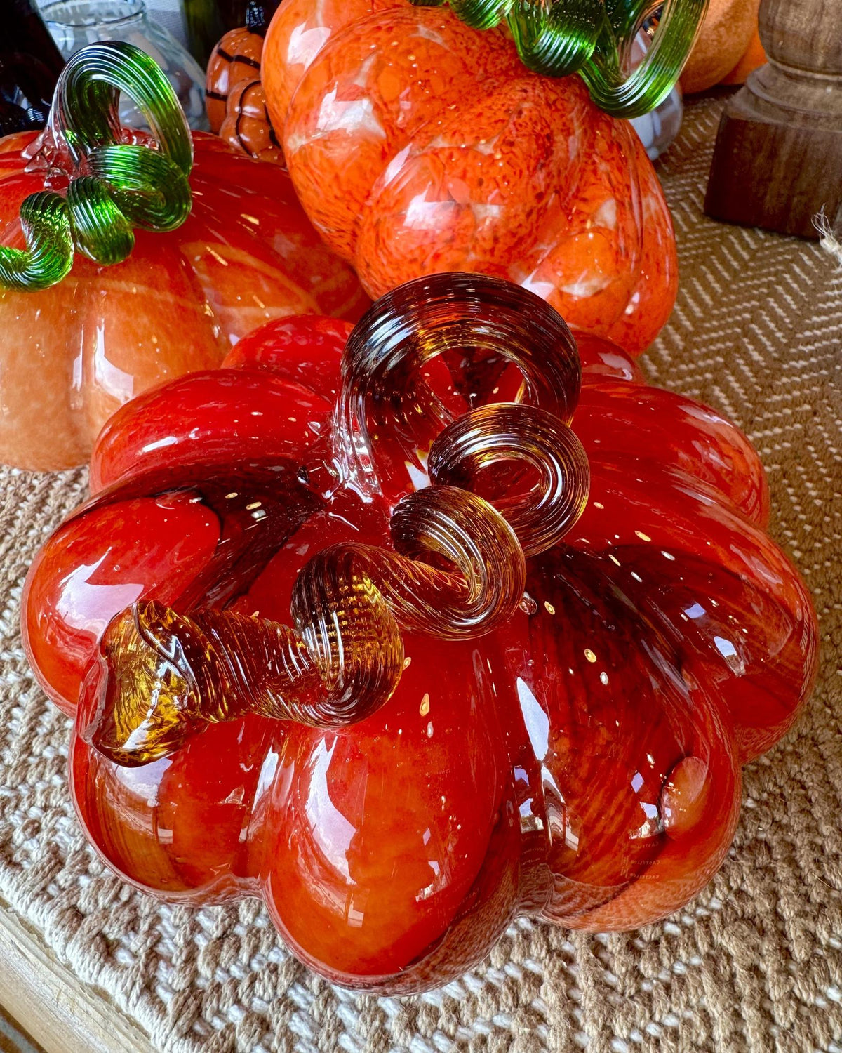 A decorative glass pumpkin sculpture in translucent orange-red tones with a curled amber stem, displayed on a woven surface with other glass pumpkins in the background.