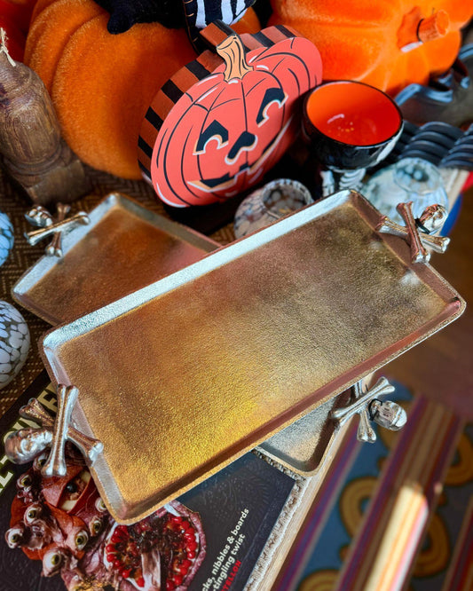 Two rectangular metal serving trays with skull and cross bone decorative handles, styled on a tabletop with Halloween decorations including pumpkins, a candle, and a book.