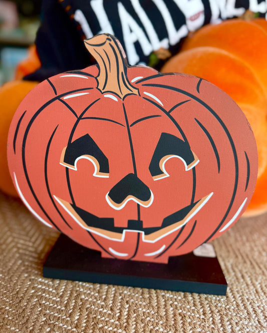 A carved jack-o'-lantern pumpkin decoration on a black base, photographed in a styled setting with pumpkins and a blurred background.