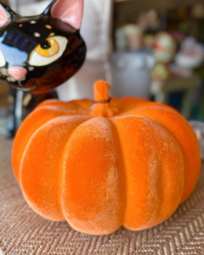 An orange pumpkin-shaped decorative object with a soft velvety texture sits on a woven beige surface. A black cat figurine is partially visible in the blurred background.