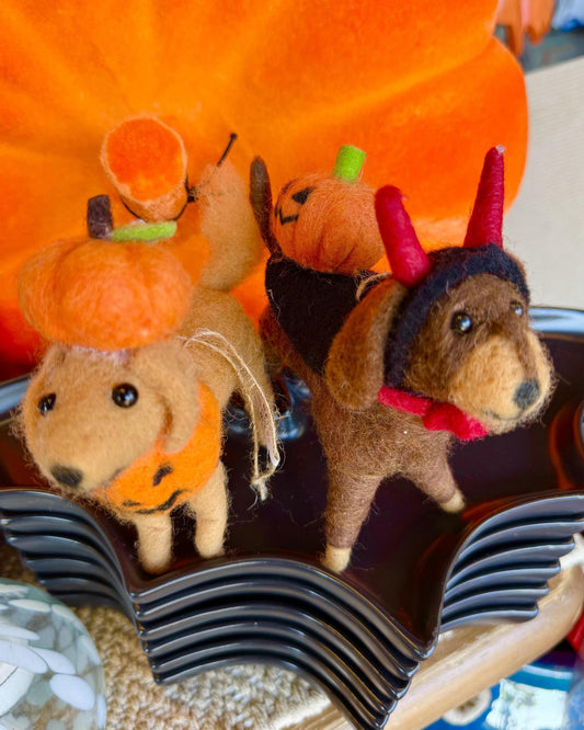 Two handmade felt dog figurines dressed in Halloween costumes, one wearing an orange pumpkin hat and the other a devil outfit with red horns and a red collar, displayed on a black wavy tray in front of a large orange pumpkin.