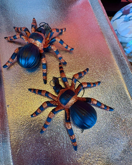 Two decorative spider ornaments with glittery copper-orange and black-and-blue striped legs, displayed on a reflective metallic surface.