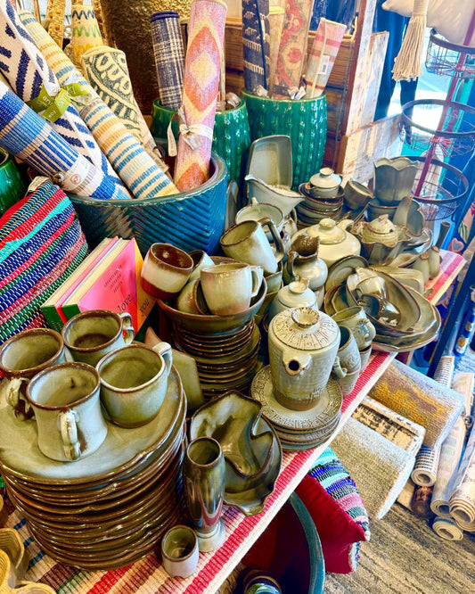 A display of Frankoma pottery tableware, including plates, mugs, cups, and small vessels, arranged on tables in an artistic shop setting with woven textiles and other home goods in the background.