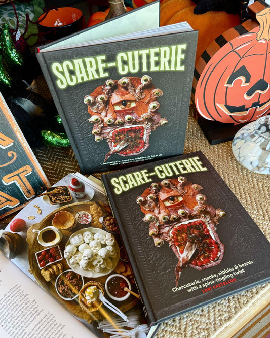 Two hardcover cookbooks titled "SCARE-CUTERIE" are arranged on a tabletop with Halloween-themed props, including a carved pumpkin, candles, and food items. The covers feature a dark horror illustration and the author name Melissa Roessler.